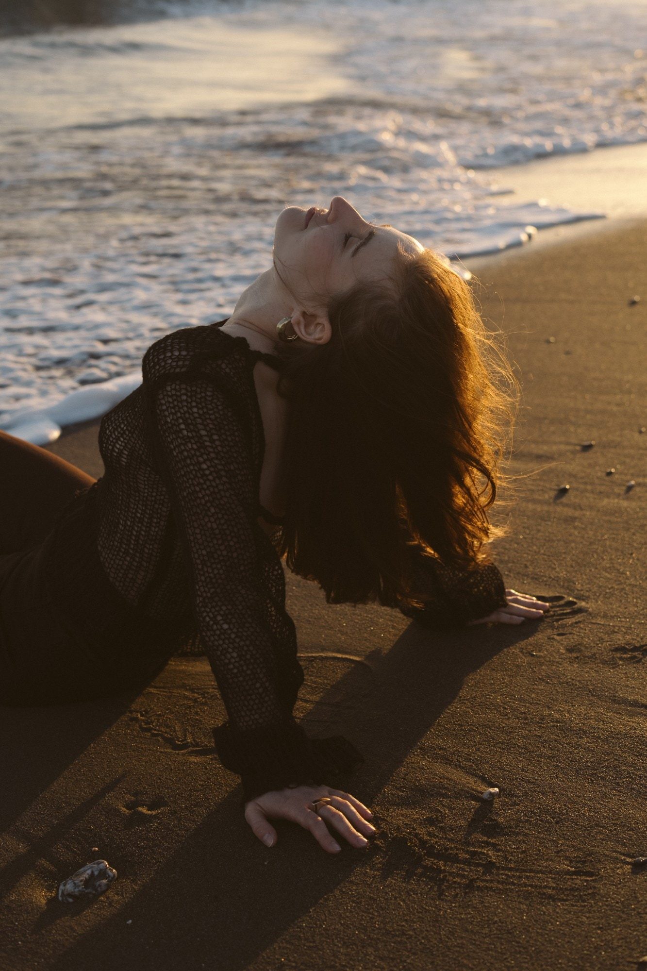 Женщина полулежит на песке у воды, запрокинув голову к закатному солнцу